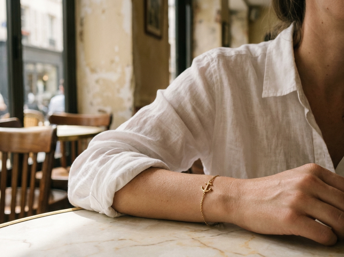 Bracelet ancre en or porté au poignet d'une femme en chemise en lin blanc dans un café parisien