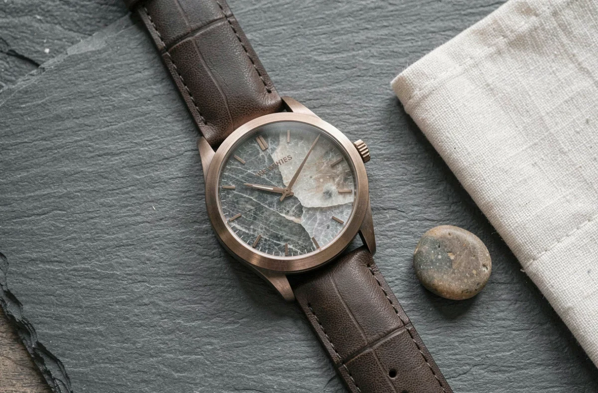 Gros plan d'une montre homme à cadran en pierre et boîtier bronze posée sur une surface en ardoise avec accessoires en lin
